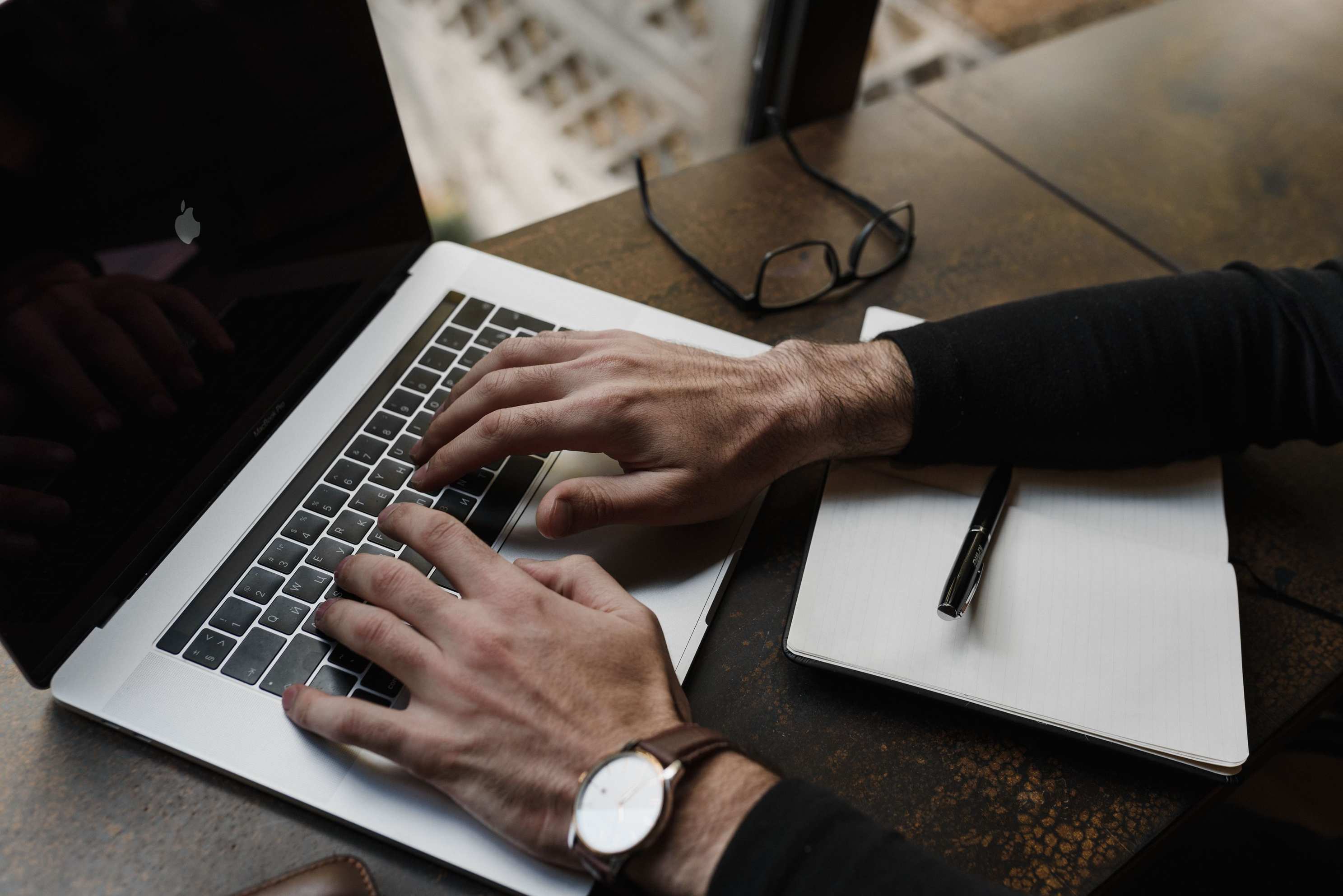 Overhead view of keyboard and notebook on a desk with hands typing on a laptop keyboard, notepad and pen nearby